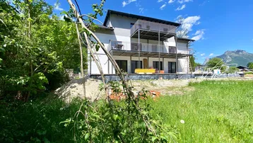 Neubaugebäude mit mehreren Balkonen in grüner Umgebung und Bergblick unter blauem Himmel.