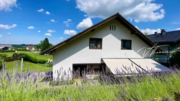 Hausfassade mit Markise und blühendem Lavendelfeld im Vordergrund, umgeben von grüner Landschaft unter blauem Himmel.