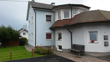 Helles Einfamilienhaus mit hellblauer Fassade, Gartenbereich und gepflastertem Vorplatz.