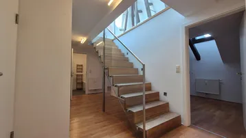 Heller Flur mit moderner Holztreppe, Glasgeländer und Parkettboden, der zu den oberen Etagen führt.