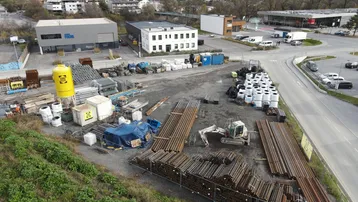 Luftaufnahme eines Gewerbegebiets mit Baumaterialien, Gebäuden und einer Straße vor einer Bergkulisse.