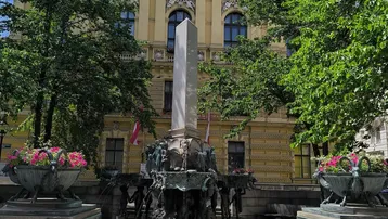 Historisches Gebäude mit klassischer Architektur, umgeben von Bäumen und einem kunstvollen Brunnen auf einem öffentlichen Platz.