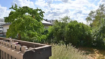 Blick vom Balkon auf den grünen Garten mit Bäumen und umliegenden Häusern unter blauem Himmel.
