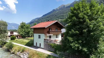 Haus am Flussufer mit Bergblick und kleiner Terrasse, umgeben von grüner Natur.