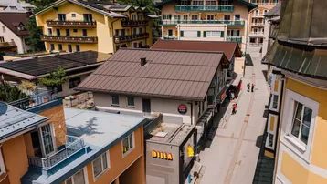 Luftaufnahme einer belebten Straße mit verschiedenen Gebäuden und Supermarkt in alpiner Umgebung.