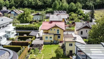 Luftaufnahme einer gepflegten Wohnsiedlung mit Häusern, Gärten und grünen Hügeln im Hintergrund.