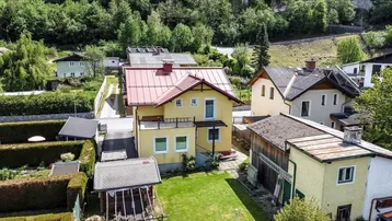 Erhöhte Ansicht des gelben Hauses mit rotem Dach, umgeben von weiteren Gebäuden und grünen Hügeln.