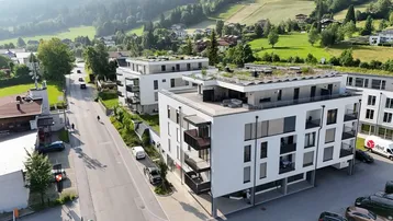 Moderne Wohngebäude mit Flachdächern und Balkonen in einer grünen, hügeligen Landschaft mit Bergblick.