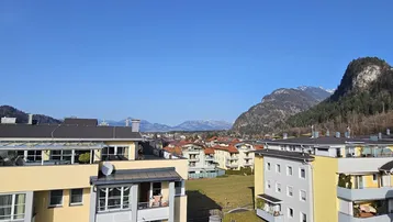 Weitläufiger Blick über die Dächer der Stadt auf die umliegende Berglandschaft unter blauem Himmel.