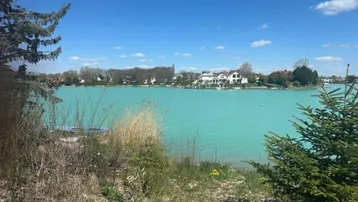 Panoramablick auf einen türkisfarbenen See mit Häusern am gegenüberliegenden Ufer unter blauem Himmel.