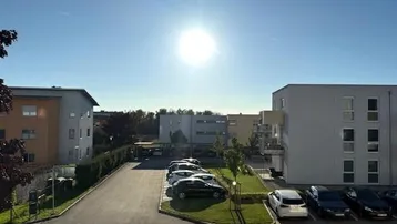 Außenansicht von Mehrfamilienhäusern mit Parkplätzen und Grünflächen unter strahlendem Sonnenschein.