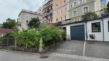 Rückansicht eines Gebäudekomplexes mit Garagenzufahrt und üppiger, wilder Vegetation.