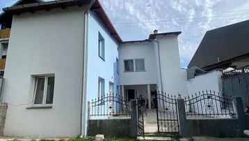 Einfamilienhaus mit hellblauer Fassade, grünem Vorgarten und schmiedeeisernem Zaun unter blauem Himmel.