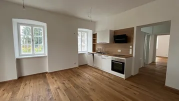 Offener Wohnbereich mit integrierter Küche, Holzboden und zwei Fenstern, bereit zur Einrichtung.
