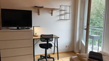 Helles Schlafzimmer mit Schreibtisch, Fernseher und Zugang zu einem Balkon.