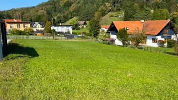 Weitläufige grüne Wiese mit Blick auf umliegende Häuser und bewaldete Hügel unter blauem Himmel.