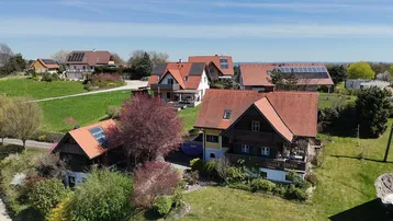 Luftaufnahme einer ländlichen Siedlung mit mehreren Häusern und grünen Gärten unter blauem Himmel.