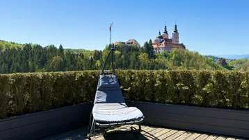 Sonniger Terrassenbereich mit Holzbelag, Liegestuhl und weitem Blick auf eine historische Kirche und grüne Hügel.