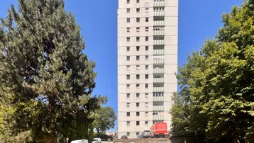 Hohes Wohngebäude mit mehreren Stockwerken, umgeben von Bäumen und Parkplätzen unter blauem Himmel.