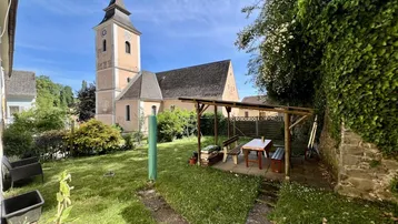 Grüner Garten mit überdachtem Sitzbereich und Blick auf die Kirche.