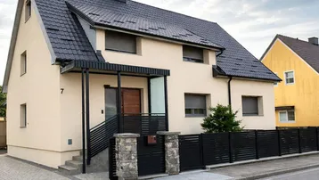Modernes Einfamilienhaus mit beiger Fassade, dunklem Dach und gepflegtem Eingangsbereich.