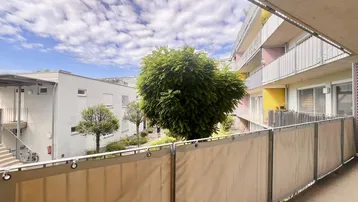 Blick vom Balkon auf den begrünten Innenhof mit weiteren Wohngebäuden und blauem Himmel.