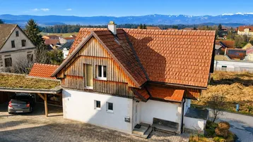 Haus mit rotem Ziegeldach, Holzfassade und Carport, umgeben von ländlicher Landschaft mit Bergblick.
