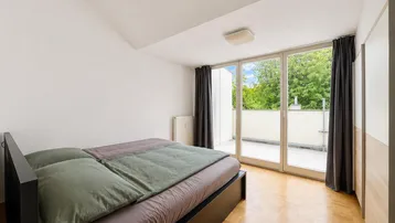 Helles Schlafzimmer mit Parkettboden, großem Bett und Zugang zu einem sonnigen Balkon.