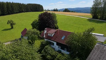 Luftaufnahme eines traditionellen Hauses mit rotem Ziegeldach, umgeben von grünen Wiesen und Wäldern.