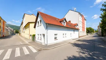 Weißes Gebäude mit rotem Ziegeldach an einer Straßenecke, umgeben von anderen Häusern und Bergen im Hintergrund.