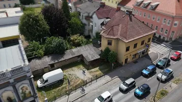 Luftaufnahme eines Gebäudes an einer Straße mit parkenden Autos und umliegender Bebauung.