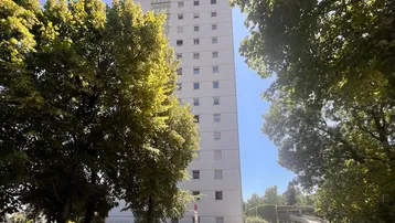 Hohes Wohngebäude mit vielen Fenstern und Balkonen, umgeben von Bäumen und Parkplätzen.