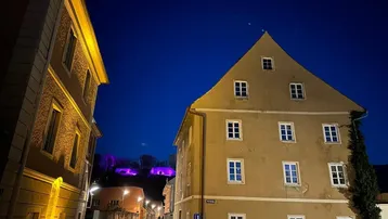 Historisches Gebäude in der Abenddämmerung mit beleuchteten Fenstern und einer lila leuchtenden Burg im Hintergrund.