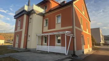Mehrstöckiges Gebäude mit orangefarbener Fassade, einer Terrasse im Erdgeschoss und einer Dachterrasse.