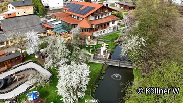 Luftaufnahme eines Anwesens mit mehreren Gebäuden, einem Teich und blühenden Bäumen in ländlicher Umgebung.