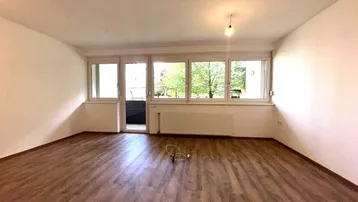 Helles, leeres Zimmer mit Laminatboden und großen Fenstern, die viel Tageslicht hereinlassen.