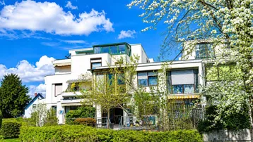 Modernes Wohngebäude mit viel Grün und blühenden Bäumen unter strahlend blauem Himmel.