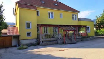 Gelbes Gebäude mit rotem Ziegeldach und überdachter Terrasse, die als Außenbereich für Gäste dient.