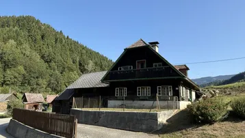 Traditionelles Holzhaus in ländlicher Umgebung mit Blick auf bewaldete Hügel unter blauem Himmel.