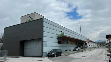 Modernes Geschäftsgebäude mit grauer Fassade und Parkplätzen an der Straße unter bewölktem Himmel.