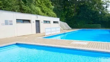 Außenbereich mit zwei Schwimmbecken und einem Funktionsgebäude, umgeben von Bäumen.