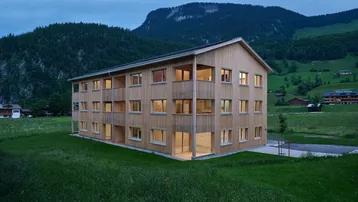 Modernes Holzgebäude in alpiner Landschaft bei Dämmerung mit beleuchteten Fenstern.