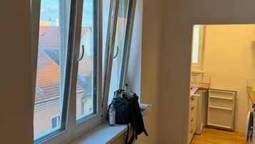 Heller Raum mit Holzboden und geöffnetem Fenster, das einen Blick über die Dächer bietet.
