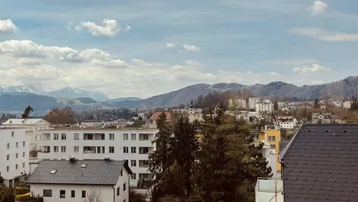 Panoramablick über eine Stadtlandschaft mit Bergen im Hintergrund unter bewölktem Himmel.
