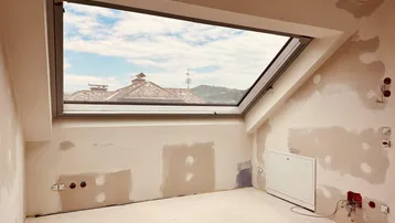 Unfertiger Innenraum mit großem Dachfenster, das einen weiten Blick auf Dächer und Hügel bietet.