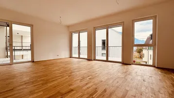 Helles, leeres Wohnzimmer mit Parkettboden und Zugang zu mehreren Balkonen mit Bergblick.
