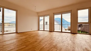 Helles, leeres Zimmer mit Parkettboden und Zugang zu mehreren Balkonen mit Seeblick.