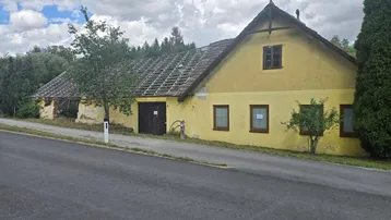 Altes gelbes Bauernhaus mit beschädigtem Dach und grüner Umgebung an einer Straße.