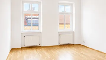 Helles Zimmer mit zwei Fenstern, Holzboden und weißen Wänden, ideal als Wohn- oder Schlafzimmer.