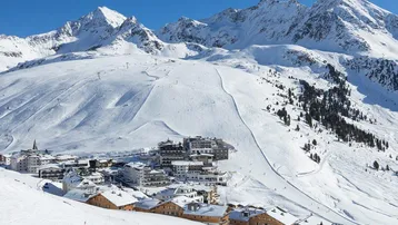 Panoramablick auf ein verschneites Bergdorf mit Skipisten und majestätischen Gipfeln unter blauem Himmel.